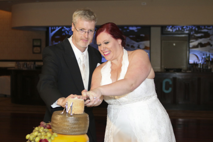 bride and groom cutting the top of the cheese wheel wedding cake